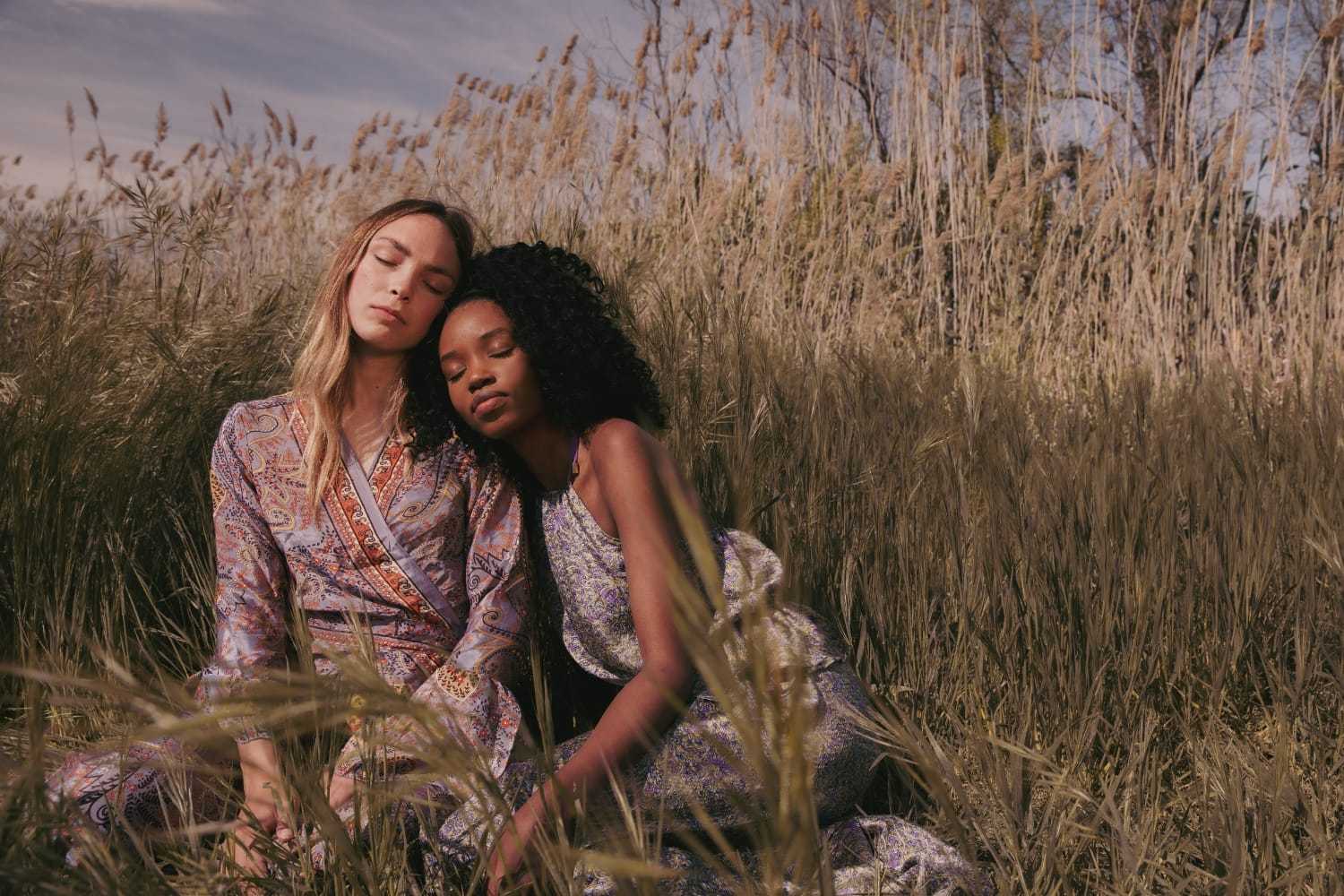 Two people sitting peacefully in a field, wearing patterned dresses amidst tall grass.