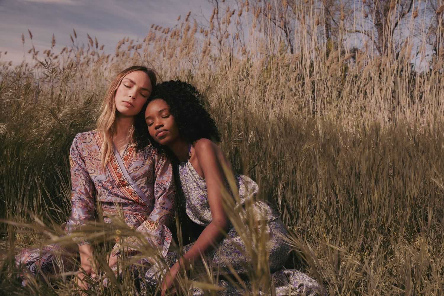 Two people sitting peacefully in a field, wearing patterned dresses amidst tall grass.